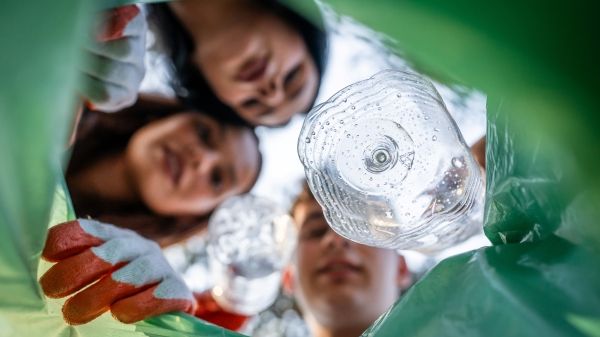 Three children look into a recycling can with a green plastic bag, putting a plastic bottle into it.