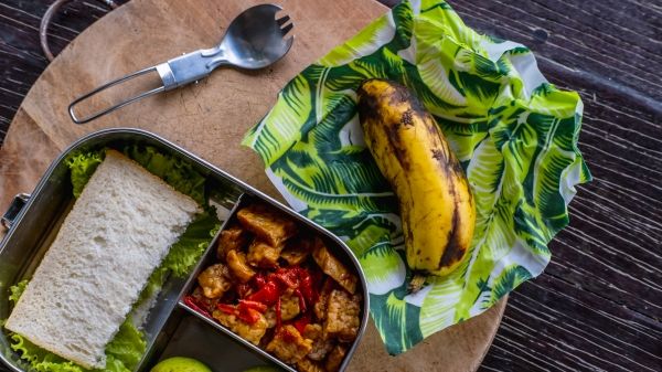 A sandwich, apples, tofu side-dish, and banana packed in an eco-friendly metal lunch box and wax paper.