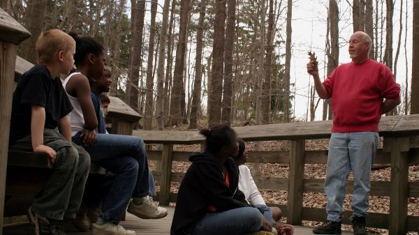 A group of people sitting on a wooden bench in a forest looking at an educator holding a leaf