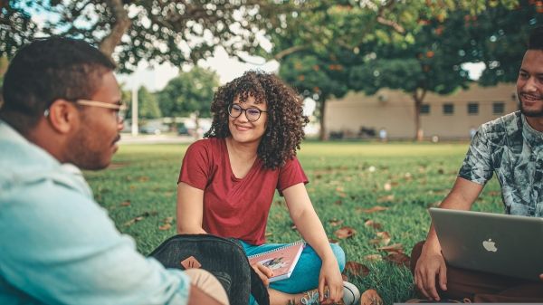 Three people sit in a grassy field talking, one with a laptop in their lap