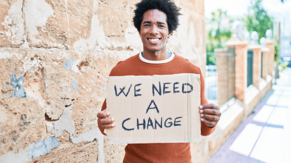 person holding "we need a change" sign on city sidewalk