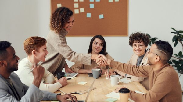 A group of six people in business casual clothes sit around a table, two reaching across the table to shake hands