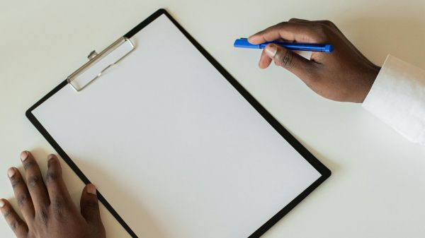 A clipboard on a white desk with two hands next to it, one holding a pen