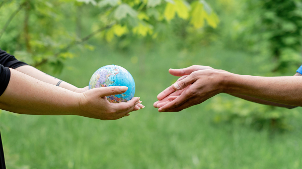 Two hands to the left pass a small globe to two hands to the right, behind them is a green plant-filled background