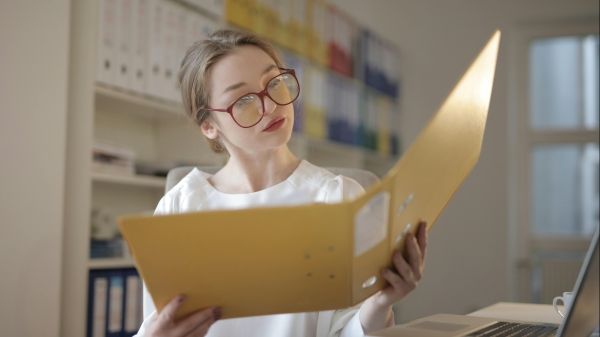 A person in red glasses and a white shirt looks at an open yellow file folder