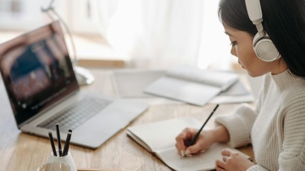 A person in front of a laptop wearing headphones and taking notes in a notebook.
