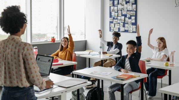 View of a teacher's back standing to the left in front of desks with students raising their hands