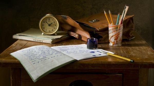 A notebook, cup full of pencils, leather bag, clock, and ink sit on a wooden desk
