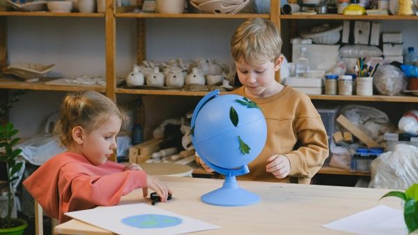 Two children interact with a globe on the table in front of them in an art classroom environment.