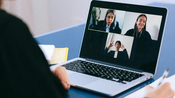 A person holding a pen over a notebook look at a laptop with a video call featuring three people