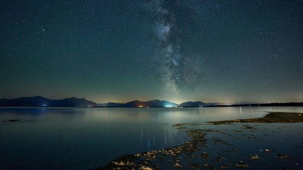 A night sky full of stars above an ocean, city lights in the distance