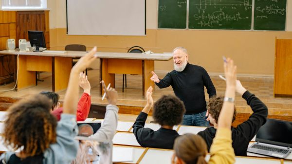 Higher Education A professor standing in front of a stage with chalkboards behind it gestures to seated students raising their hands
