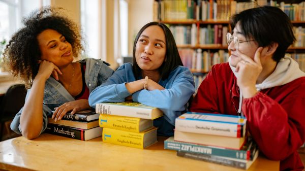 Three people sit at a table with textbooks stacked in front of them and a bookshelf behind them