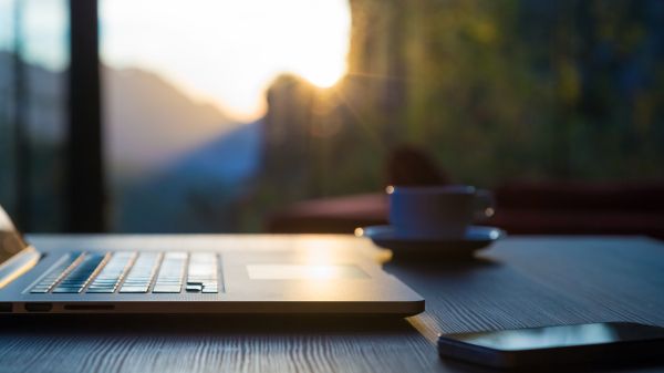 Laptop, mug, and phone lay on black wood table as sunlight shines through large windows