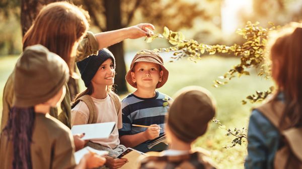 Environmental Educator A group of children are crowded around a central adult who is reaching out to touch the branch of a leafy plant