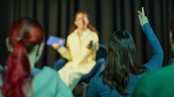 Teenage girl with brown hair sitting on stage holding tablet and remote, speaking to audience while teenage girl with dark hair raising hand in foreground, conference setting