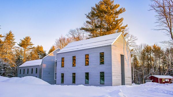A light-gray building with an A-frame roof stands prominently in morning sunlight. A layer of snow sits on the roof and deep snow is on the ground. Behind the building are leafless trees and conifers, lit brightly by yellow morning sunlight. Above the trees is blue sky.