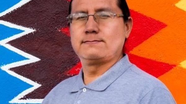 Profile photo of a man wearing glasses, hair pulled back, and staring straight at the camera. He stands in front of a colorful, geometric backdrop