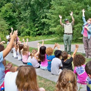 A group of people with their arms outstretched toward the sky, sitting in an open park space, while two adults are standing in front of the group