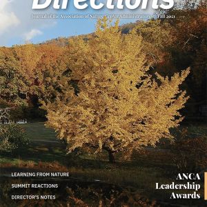 A magazine cover features a tree with bright yellow-orange leaves. A body of water at the bottom partially reflects the tree’s color. A hillside of trees is in the background. The top of the cover reads “Directions, Journal of the Association of Nature Center Administrators / Fall 2021.”