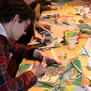 A teacher making art at a table with others and many scattered art supplies