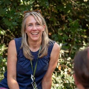 A long-haired person smiles widely at two people in the foreground. Behind them, foliage speckled with sunlight.