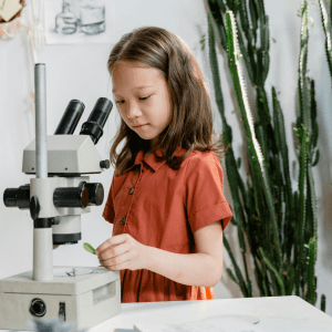 A child stands behind a microscope with a leaf in their hand; behind them is a wall featuring a skeleton diagram