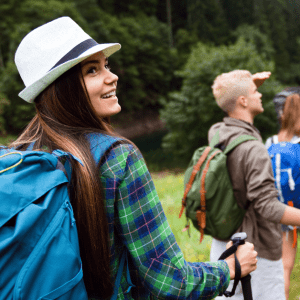 A group of young hikers wearing backpacks and holding trekking poles walk along a grassy trail near a forest and lake