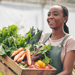 A person inside a greenhouse holds a crate full of vegetables including carrots, lettuce, and peppers