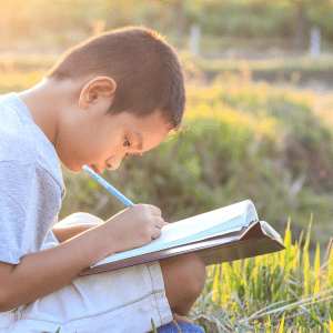 A child sits in the grass writing in a notebook; behind them is a setting sun