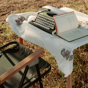 A green folding chair sits to the left of a table with a white cloth on it; on top of the table is a green typewriter and open book; behind this the ground is covered with yellow grass