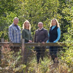 Four light-skinned people stand on a boardwalk with a wooden rail. Surrounding them is green foliage from herbaceous plants, shrubs, and trees.