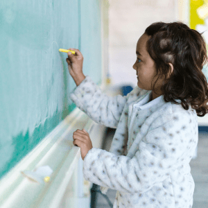 A child writes on a green chalkboard with a piece of chalk held in their right hand; behind them is a classroom wall