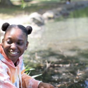 A smiling Black woman looks off to the side while near a duck pond