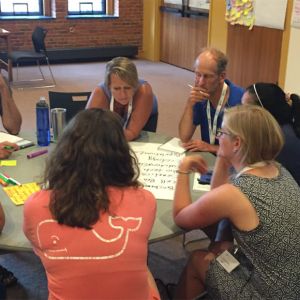 Group of adults with thoughtful expressions gathered around a round table with markers and chart paper. 