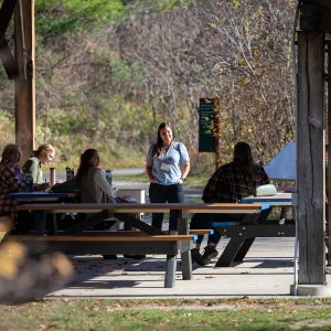 A group of people at picnic tables underneath a large outdoor shelter. In the middle, one person in a light-blue shirt stands and faces the others. In the background, mostly leafless trees with a few green conifers.