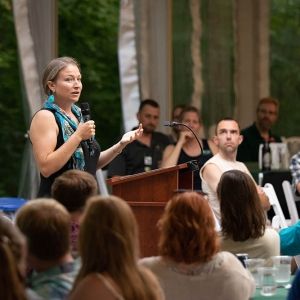 A person stands at a podium and holds a microphone, addressing a crowd. In the foreground and background, the seated crowd listens.