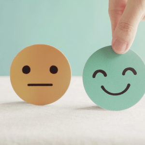 Three circles with simple faces on them sit in front of light green and white background: a red circle with a frowning face, a yellow circle with a neutral face, and a green circle with a smiling face; the green circle is being picked up by a hand