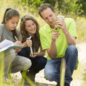 Three people squat near a creek to their right; the person on the very right holds a jar of water that they are pointing to, the person on the very left is writing in a notebook