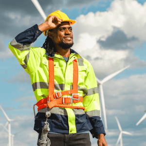 A person in a yellow visibility shirt to the left has a hand up to their hardhat; behind them is a cloudy sky with a series of wind turbines in front of it
