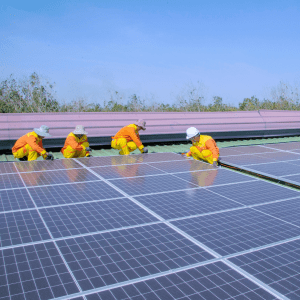 Four people in high-visibility wear kneel behind a solar panel array; behind them are trees and a blue sky
