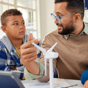 A teacher sits at a table surrounded by young students, pointing to a miniature wind turbine
