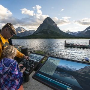 An adult an a child read an interpretive sign next to a mountain range and a body of water