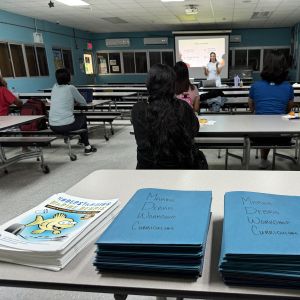 A presenter is speaking to a classroom of people with a stack of marine debris curriculum workbooks piled on one of the desks. 