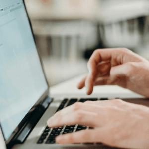 Two hands type on a laptop on a white table; white chairs can be seen in the blurred background