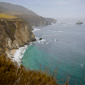 Coastal cliffs to the left of a blue ocean, a slightly cloudy blue skey behind them