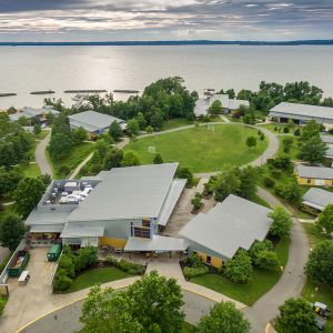 An aerial view of a campus with many gray-roofed buildings; trees and paths are interspersed between the buildings. In the background, a large body of water.