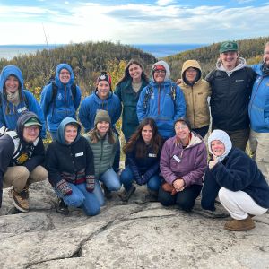A group of Wolf Ridge naturalists stand atop a rocky outcrop overlooking forested bluffs and Lake Superior.