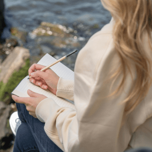 Rocks with algae sit in water to the left; to the right a person is sitting, holding a journal in their lap with a pencil in their right hand