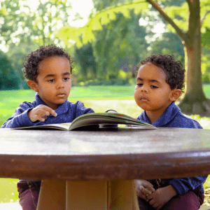 Two children sit at a wooden table with a book in front of them and a grassy field and trees behind them